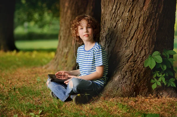 The boy of 8-9 years sits in park under a tree with the tablet in hands.