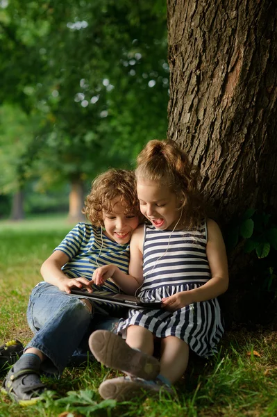 Brother with sister of 7-9 years sit under a tree and look in the tablet.