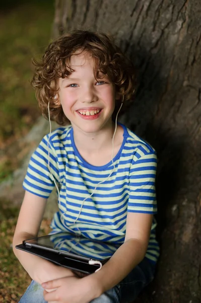 The cheerful boy sits under a tree with tablet on lap.