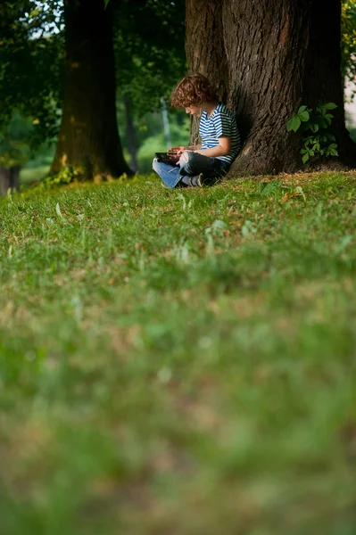 The boy sits having crossed legs near a big tree in park.