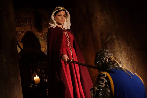 Medieval queen doing knighting ceremony in ancient castle interior.