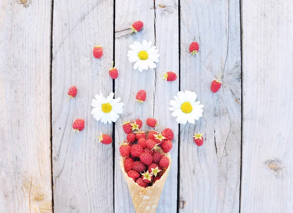 Ripe fresh raspberries and white chamomile flowers in ice cream cone on rustic wooden background. Stylish flat lay. Minimal concept.