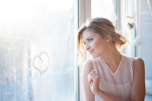 Beautiful woman with fresh daily makeup and romantic wavy hairstyle, sitting at the windowsill, draws on glass
