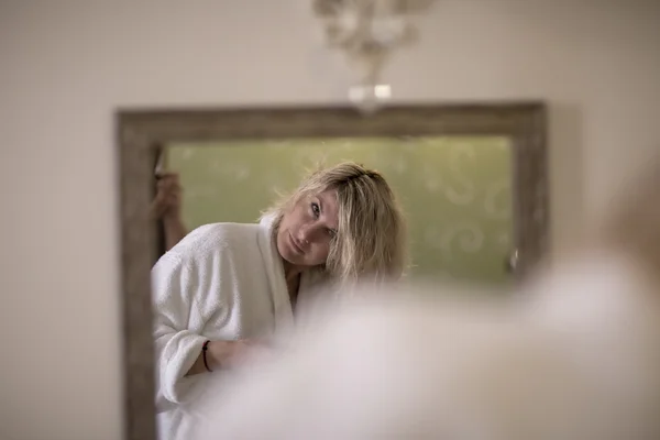 Beautiful blondie woman in the bathrobe is looking at herself in the mirror