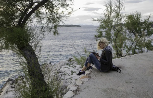 Beautiful blonde girl is seating on the stones near of the sea and watching the tablet