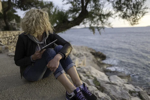 Beautiful blonde girl with curly hair is seating on the stones near of the sea with tablet in hands and watching into the distance.