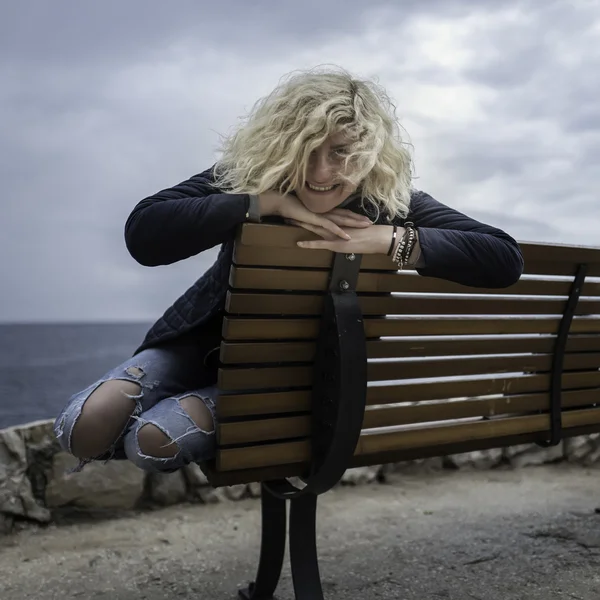 Beautiful blonde girl with curly hair in ripped jeans is seating on the bench near of the sea