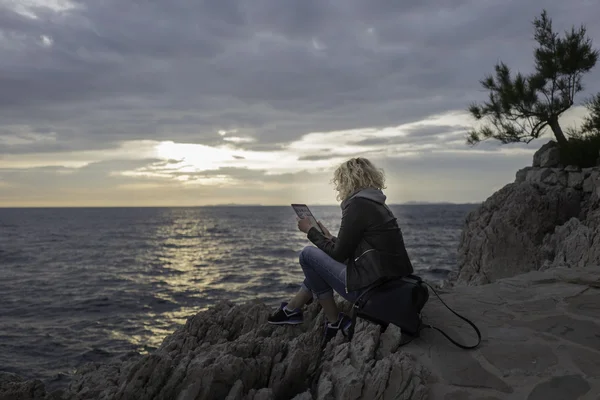 Beautiful blonde girl is seating on the stones near of the sea and watching the tablet