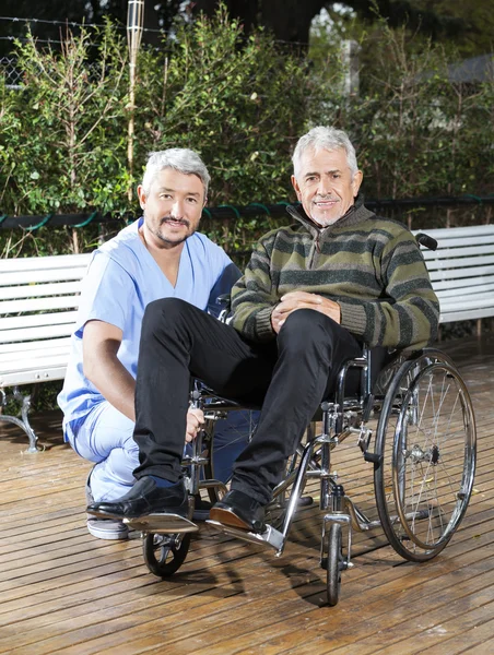 Physiotherapist Crouching By Senior Man In Wheelchair At Lawn