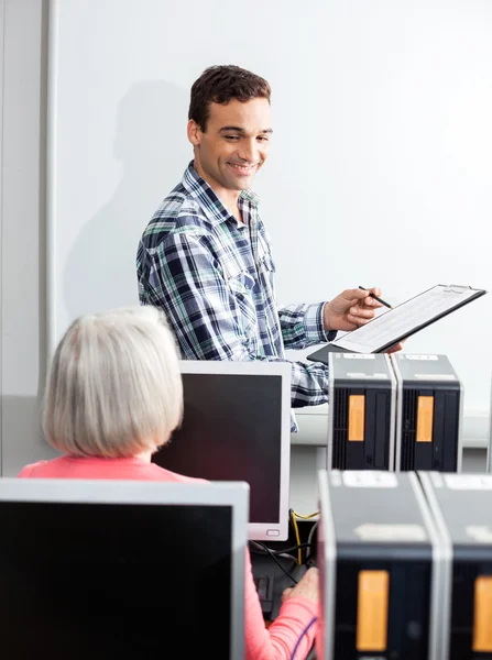 Happy Young Tutor Holding Clipboard In Computer Class