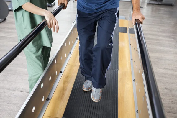 Senior Man Walking With Support Of Bars By Female Physiotherapis