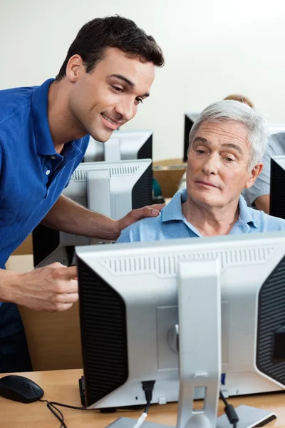 Tutor Assisting Senior Man In Using Computer At Classroom