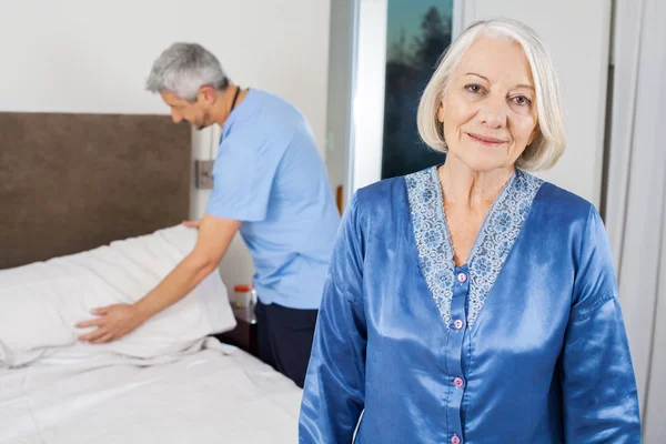 Senior Woman With Caretaker Making Bed At Nursing Home
