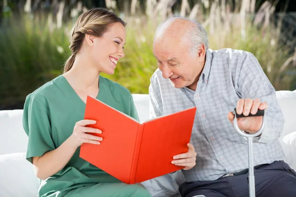Smiling Female Nurse Looking At Senior Man While Reading Book