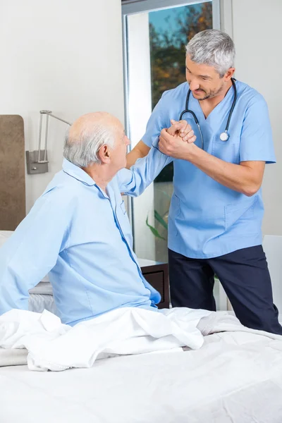 Caretaker Supporting Senior Man To Get Up From Bed