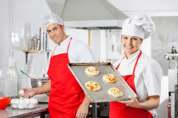 Smiling Chef Holding Pizzas On Tray With Colleague In Background