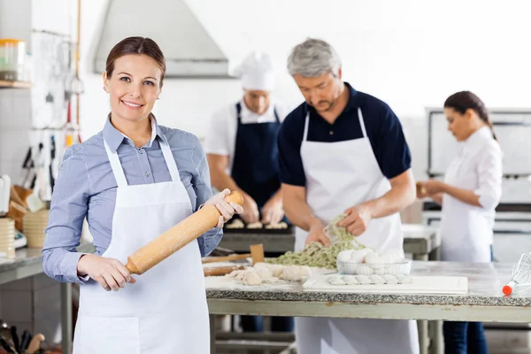 Confident Female Chef Holding Rolling Pin While Colleagues Prepa