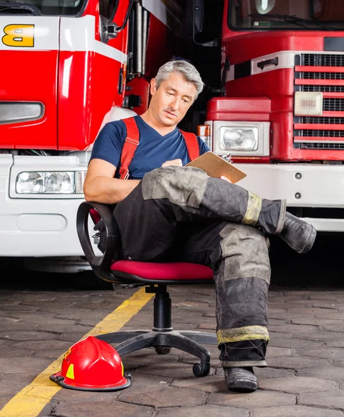 Fireman Writing On Clipboard While Sitting Against Trucks