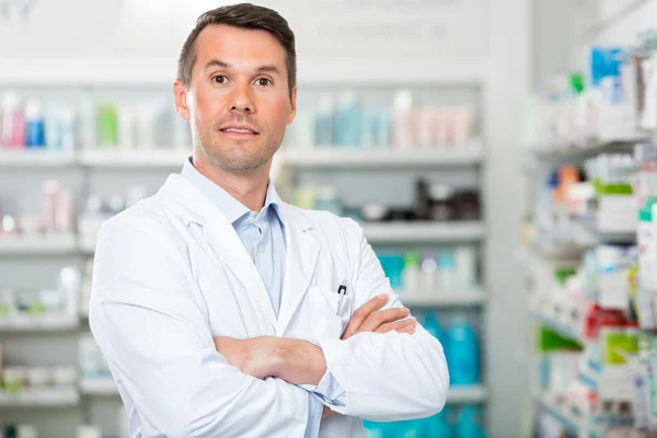 Male Pharmacist With Arms Crossed Standing In Pharmacy