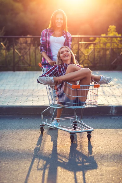 Two happy beautiful teen girls driving shopping cart outdoors