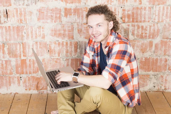 Happy smiling man typing on laptop in front of a brick wall. Happy hipster working in room. Modern lifestyle