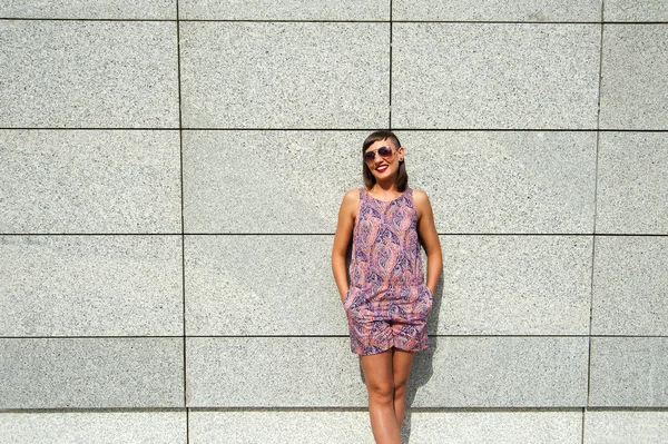 Young modern woman in sunglasses by the wall in city smiling at
