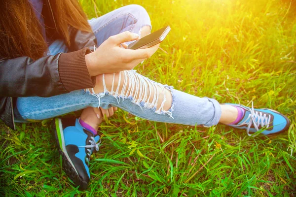 Young girl in leather jacket and ripped jeans sitting on the grass in the park and talking to friends on the phone.