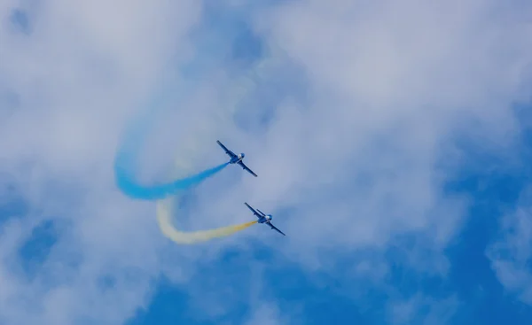 Two aircraft blue during a performance at an airshow stunt produce smoke strip of blue and yellow in the sky against a background of clouds.