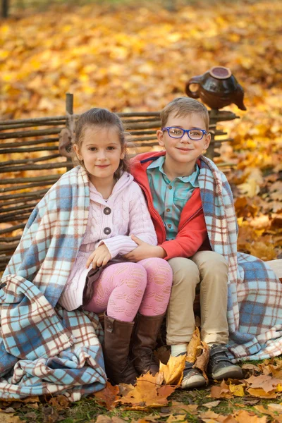 Two friends: a boy and a girl in autumn park sitting on wooden bench near a fence