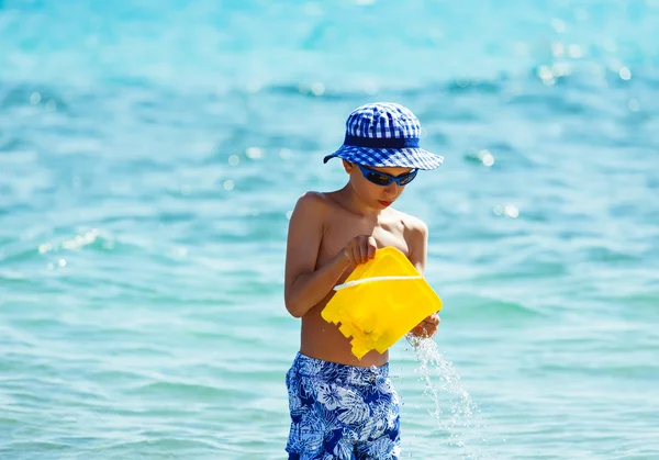 Funny little boy with a panama playing in sea splashing water with toy bucket