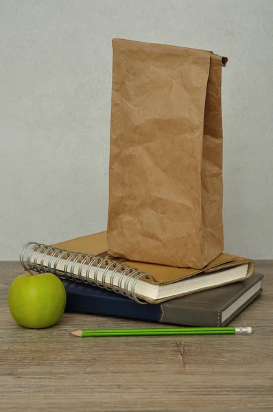 A green apple. a paper lunch bag and a stack of books on a table