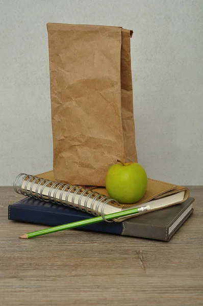 A green apple. a paper lunch bag and a stack of books on a table