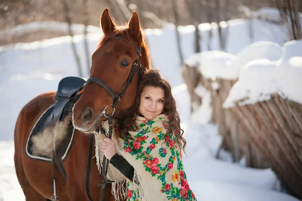 Beautiful young woman in Russian national scarf with a horse in the countryside in winter