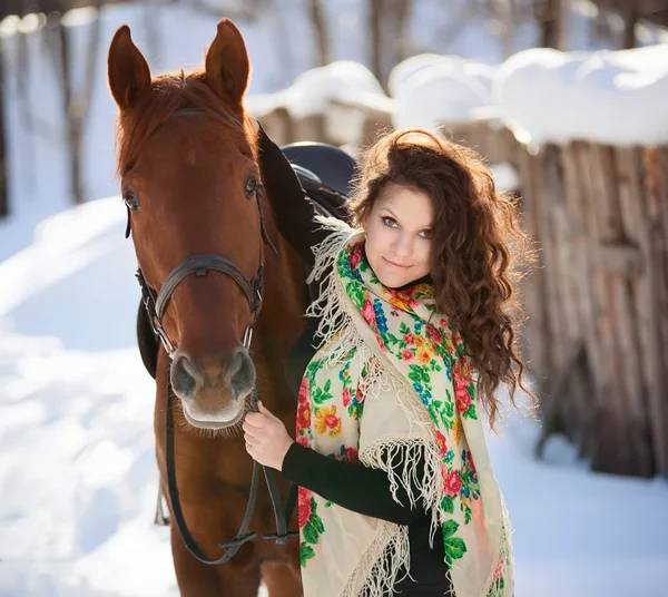 Beautiful young woman in Russian national scarf with a horse in the countryside in winter