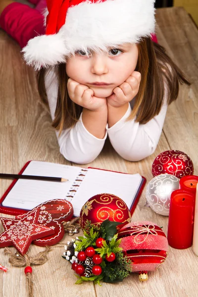 Funny girl in Santa hat writes letter to Santa near christmas decoration