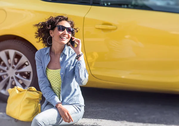 Young woman sitting next to yellow car and talking on a mobile p