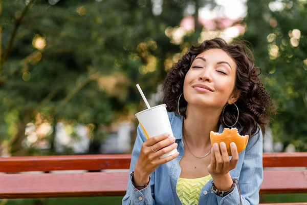 Happy woman on the bench in park eating a fast food hamburger an