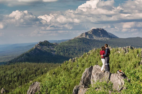 Rear view, couple hikers looking into the distance, is standing on the top of mountain rock. Background is mountain range and blue sky.