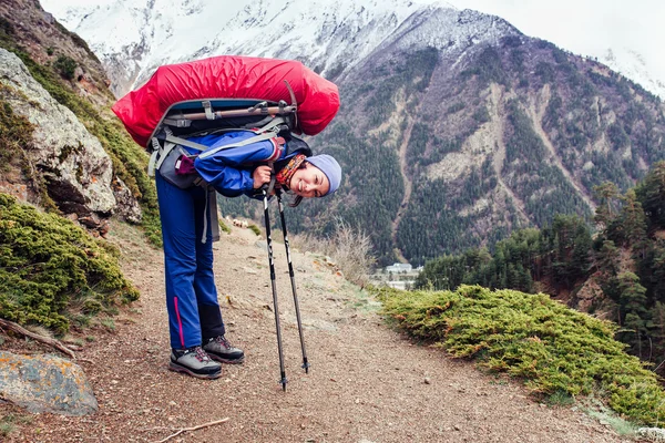 Hiking woman with backpack have a rest on a trail with her trekking poles on the background of high Caucasus mountains