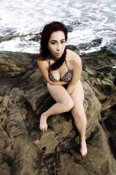 Latina Woman Sitting On Rocks In Bikini With Ocean Behind Her