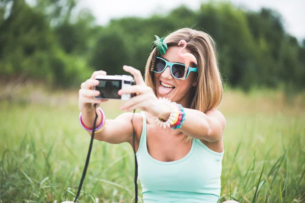 Young woman taking self portrait outdoor, wearing funny sunglasses and holding a compact camera