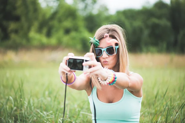 Young woman taking self portrait outdoor, wearing funny sunglasses and holding a compact camera