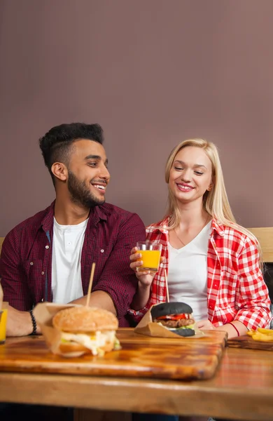 Young Woman And Hispanic Man Sitting In Cafe Hold Orange Juice Order Beef Burger Served In Paper On Wooden Table