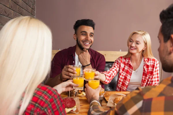 Young People Group Man And Woman Sitting In Burger Cafe, Toasting Orange Juice Order Fast Food On Wooden Table