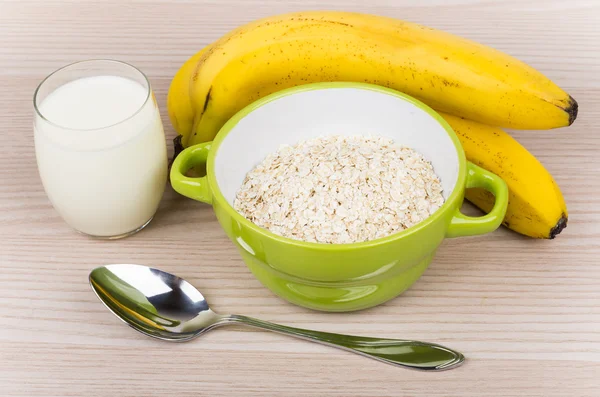 Dry oat flakes in green bowl, glass of milk, bananas