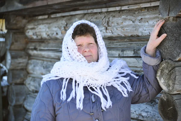 The Russian woman in  shawl warms hands near an izba