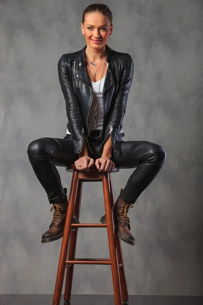 Blonde in leather and boots, posing seated on stool in studio
