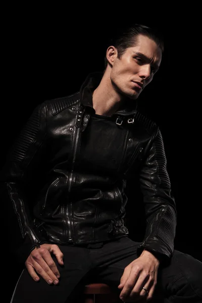 rocker in leather jacket posing seated in dark studio backgroun