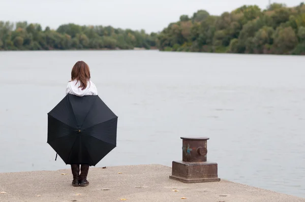 Lonely teenage girl with umbrella standing on river dock on gloomy autumn day