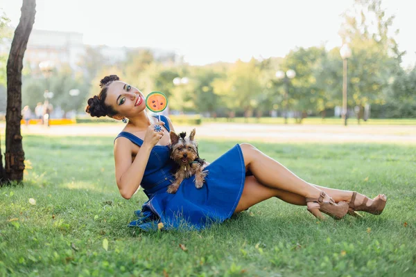 Bright makeup beautiful girl with Yorkshire Terrier seating on the grass holding watermelon lollipop.